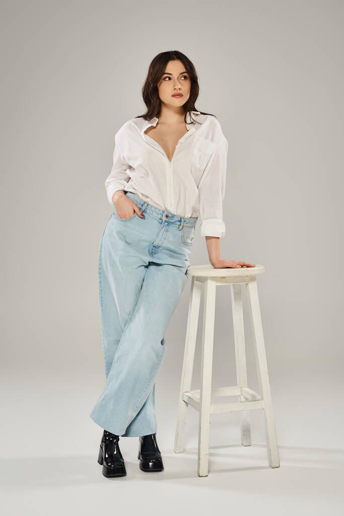 Красива жінка плюс розмір у білій сорочці і джинсах позує витончено, коли вона спирається на табурет на сірий фон. - Фото, зображення