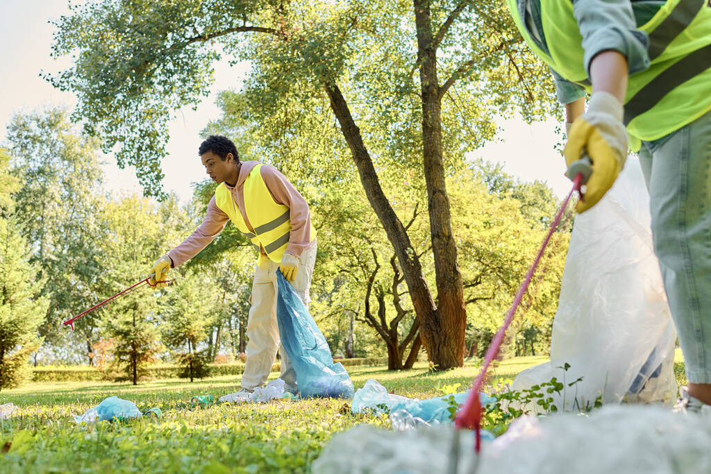 Una pareja diversa, con chalecos y guantes de seguridad, trabajando juntos para limpiar un parque cubierto de hierba. - Foto, Imagen