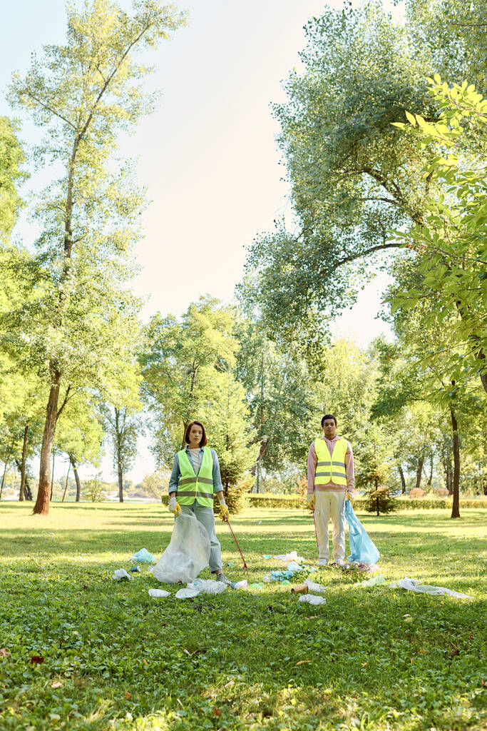 Aktywna społecznie, zróżnicowana kochająca się para w kamizelkach i rękawiczkach, stojąca na trawie, czyszcząca razem park. - Zdjęcie, obraz
