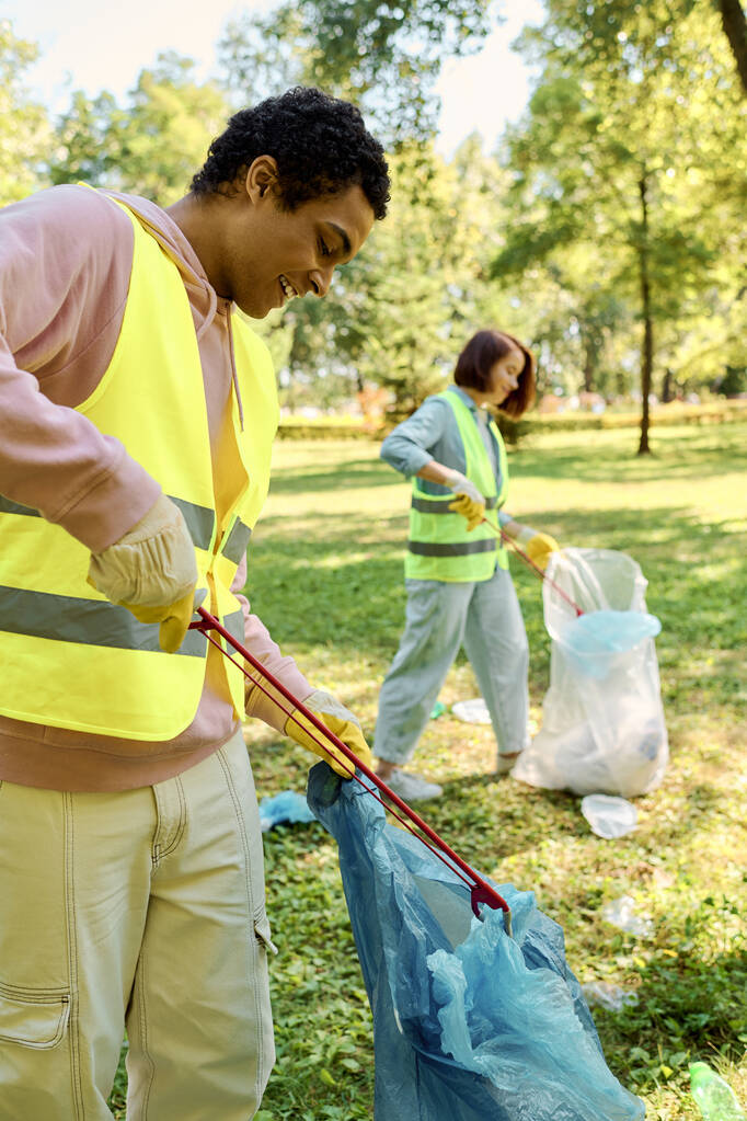 Socially active, diverse couple in safety vests and gloves cleaning the park together with trash bags. - Photo, Image