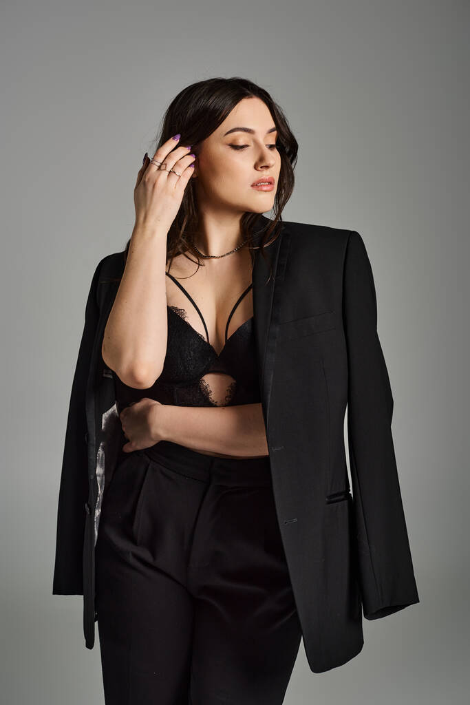 Een mooie plus size vrouw gekleed in een zwart pak slaat een zelfverzekerde pose tegen een grijze achtergrond. - Foto, afbeelding