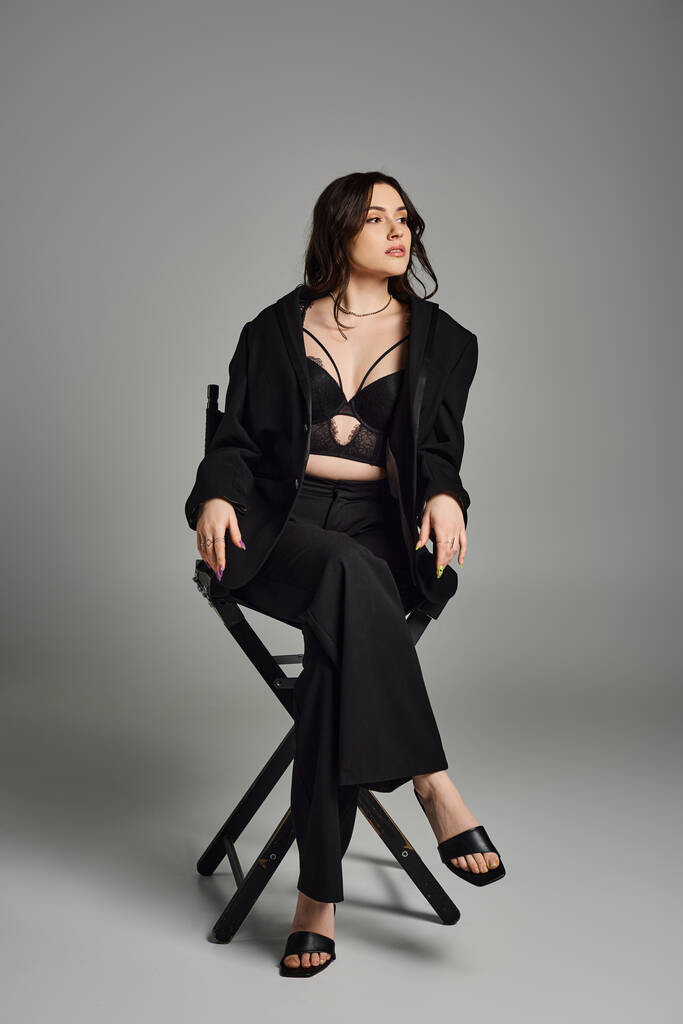 Uma mulher plus size em traje elegante sentado em uma cadeira, cruzando as pernas graciosamente em um pano de fundo cinza. - Foto, Imagem