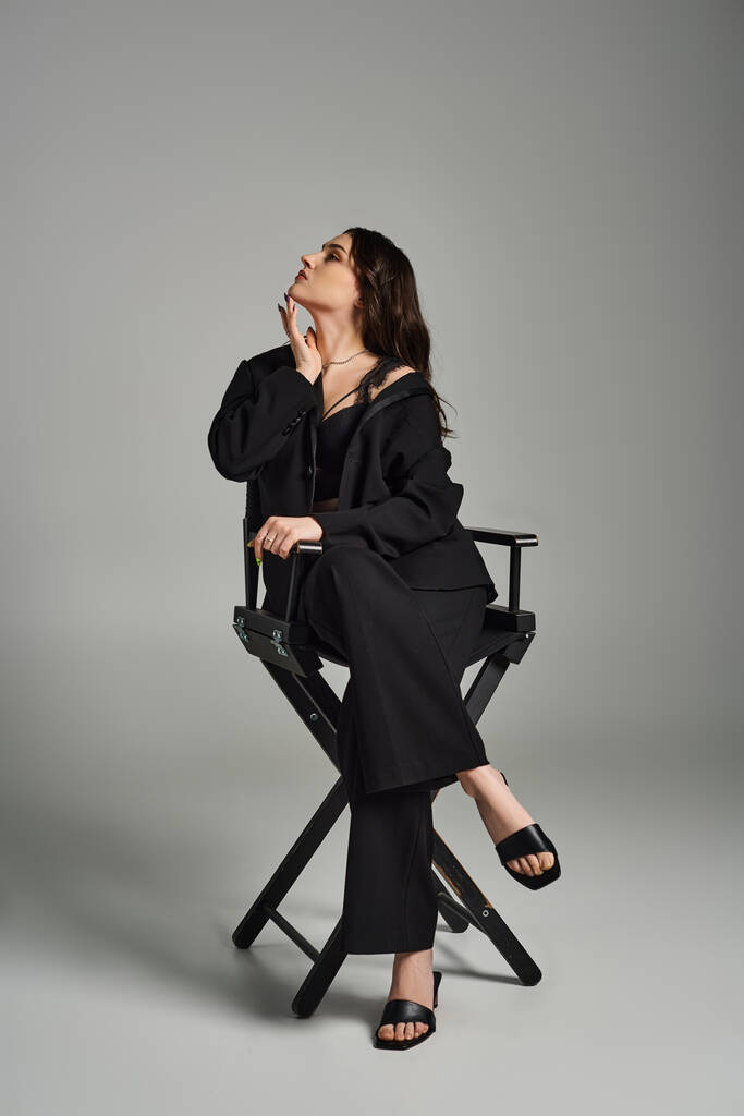 Una hermosa mujer de talla grande con un vestido negro se sienta con gracia en una silla contra un fondo gris, exudando confianza. - Foto, imagen