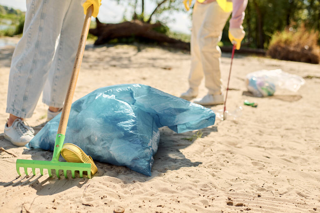 Een sociaal actief, divers liefdevol koppel, met veiligheidsvesten en handschoenen aan, staand op het strand met een schop en een zak vuilnis. - Foto, afbeelding