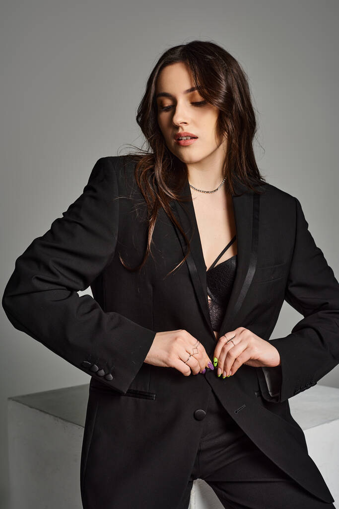 Uma bela mulher plus size confiantemente posa em um terno preto e amarra contra um pano de fundo cinza. - Foto, Imagem