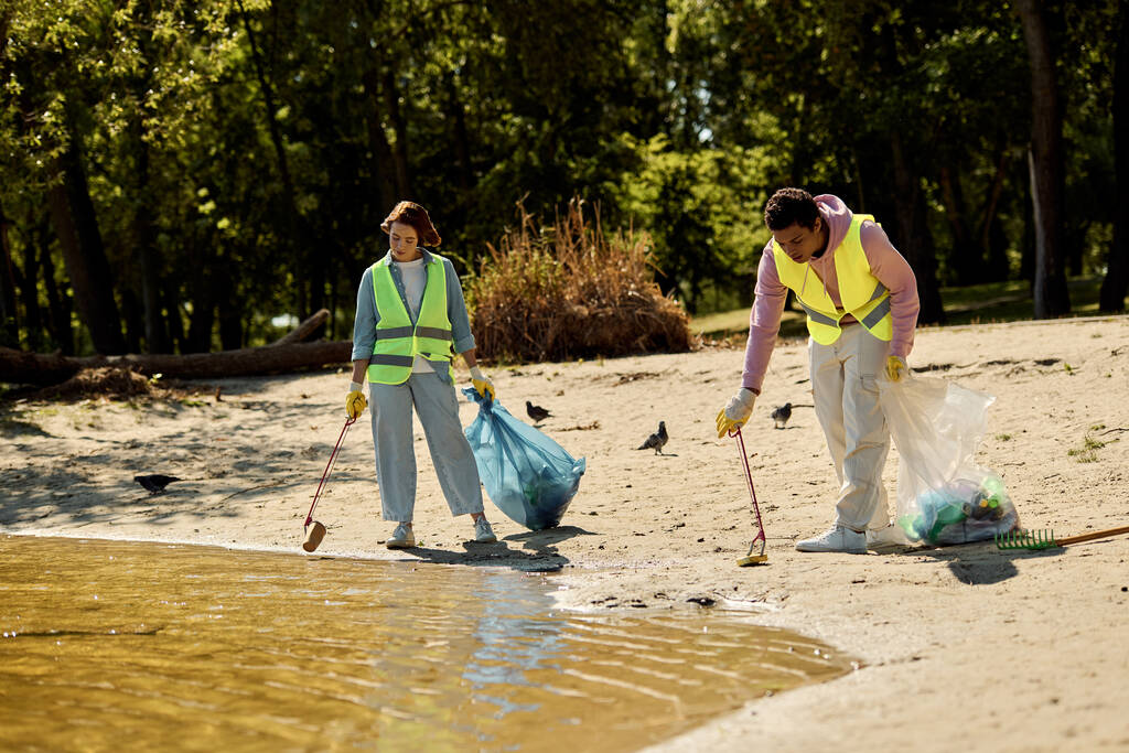 Un couple aimant et socialement actif, portant des gilets de sécurité et des gants, nettoie le parc ensemble tout en restant debout dans le sable. - Photo, image
