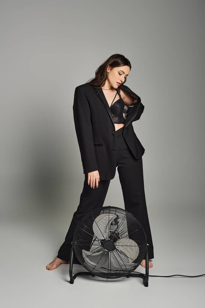 Стильна жінка плюс розмір в костюмі, впевнено стоїть поруч з вентилятором на сірому тлі, виключаючи зіткнення і грацію. - Фото, зображення