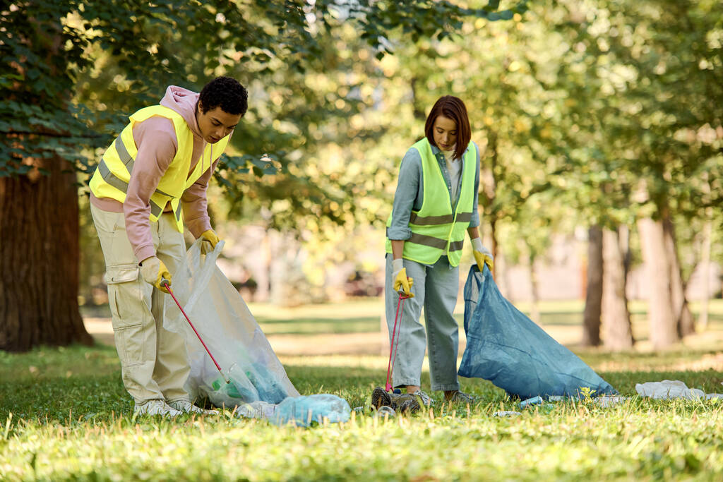 Un couple diversifié et aimant, vêtu de gilets de sécurité et de gants, debout dans l'herbe verte pendant qu'ils nettoient le parc ensemble. - Photo, image