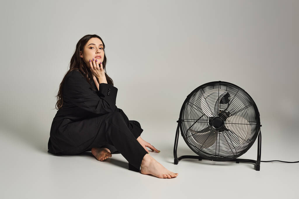 Egy gyönyörű, plusz méretű nő elegáns öltözékben, aki kecsesen ül a padlón egy ventilátor mellett, szürke háttér mellett.. - Fotó, kép