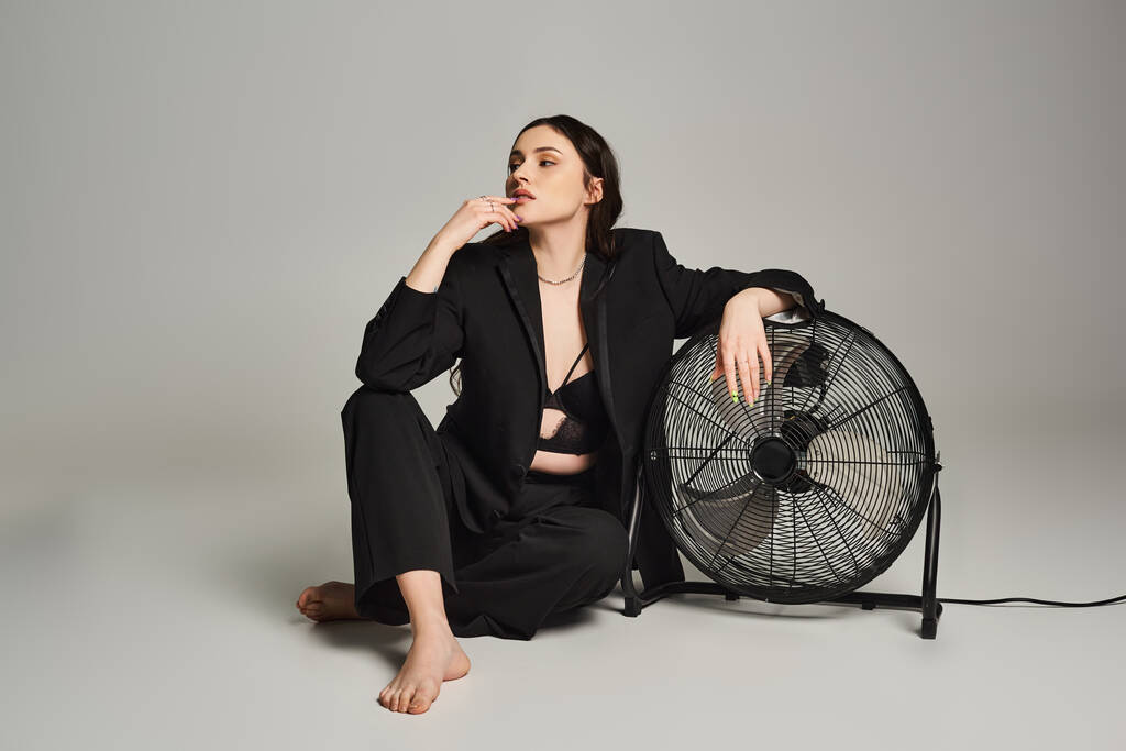 Plus velikost žena ve stylovém oblečení sedí elegantně vedle ventilátoru, objímající okamžik klidu a klidu. - Fotografie, Obrázek