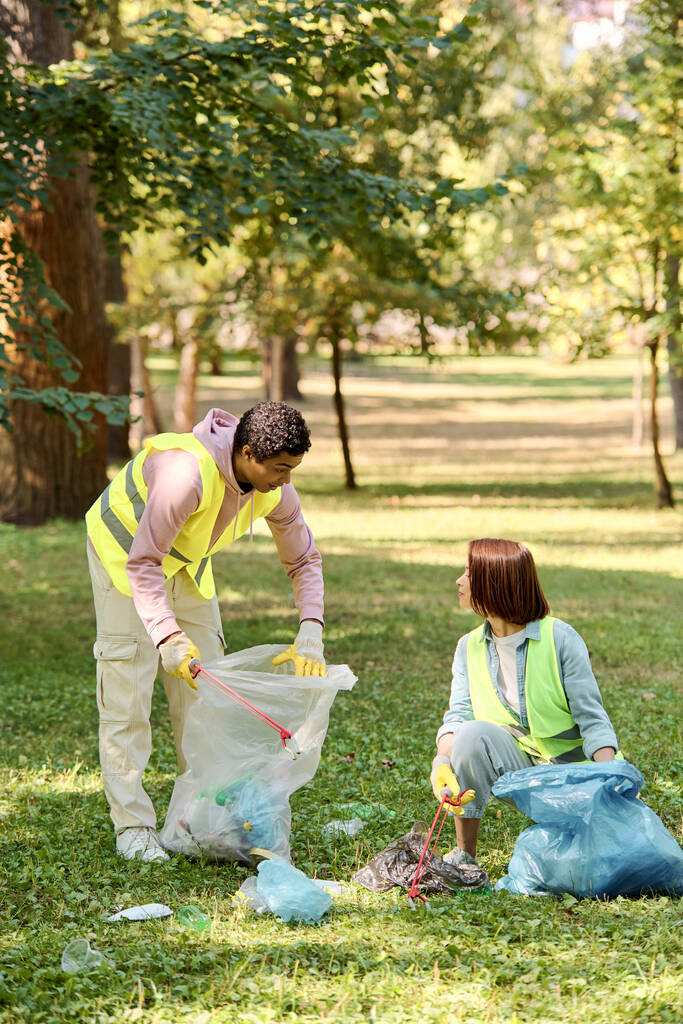 Африканский американец и кавказка в защитных жилетах и перчатках работают вместе, чтобы собирать мусор в парке, содействуя экологичности и уходу за обществом. - Фото, изображение