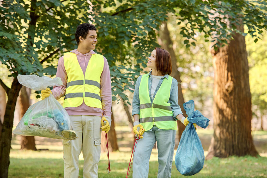 Una pareja socialmente activa y diversa en chalecos de seguridad y guantes limpiando un parque juntos, de pie en la exuberante hierba verde. - Foto, imagen
