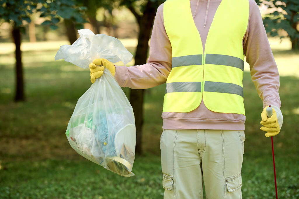 Αφροαμερικάνος με ένα φωτεινό κίτρινο γιλέκο ασφαλείας κρατάει μια σακούλα σκουπίδια ενώ συμμετέχει σε μια δραστηριότητα καθαρισμού πάρκου.. - Φωτογραφία, εικόνα