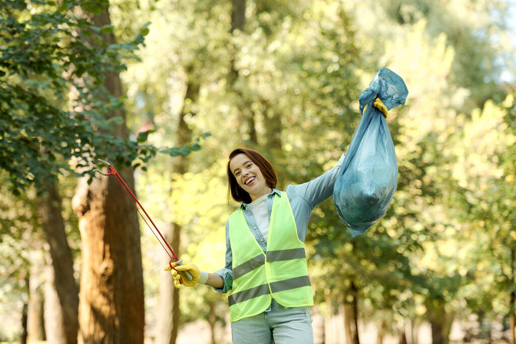 A woman in a bright safety vest is holding a vibrant blue bag while cleaning up a park with her partner in the background. - Photo, Image