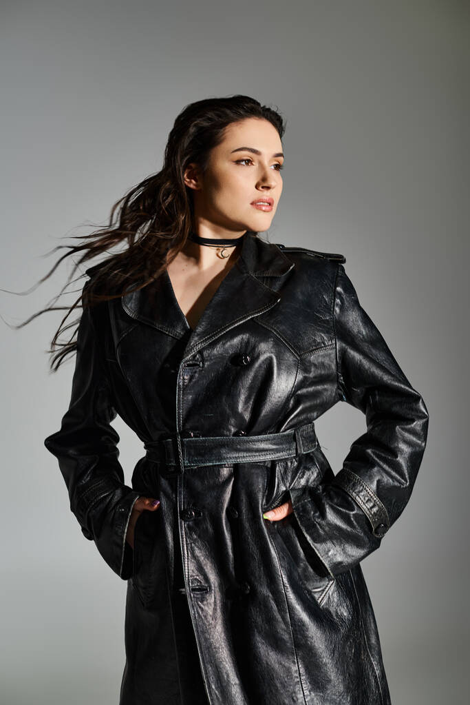 A beautiful plus-size woman poses gracefully in a chic black leather trench coat against a neutral gray backdrop. - Photo, Image