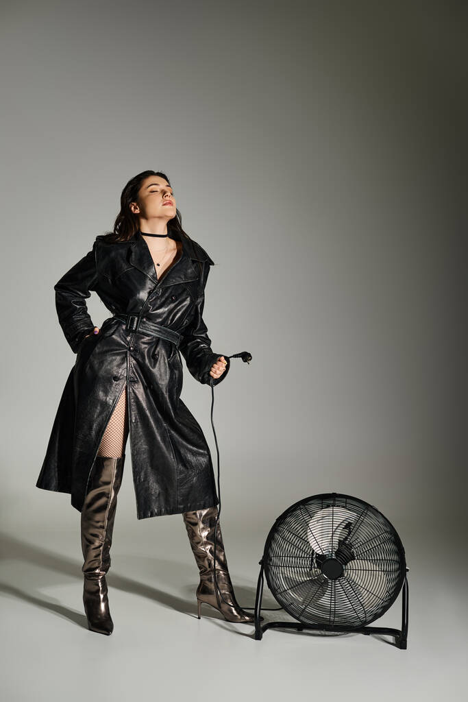 Hermosa mujer de talla grande en un abrigo negro de pie con gracia junto a un ventilador en un fondo gris. - Foto, imagen