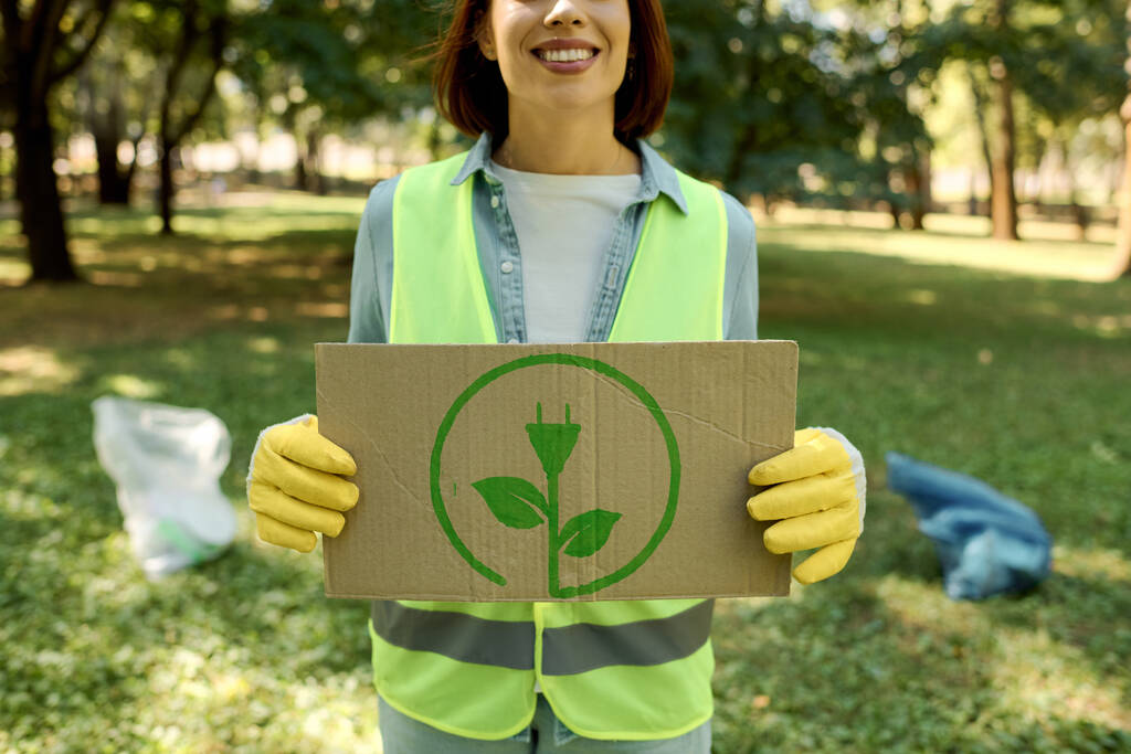 Una donna in gilet di sicurezza tiene con attenzione una scatola di cartone con una pianta verde su di esso mentre si impegna in giardinaggio sostenibile. - Foto, immagini