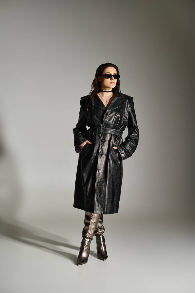 Μια όμορφη γυναίκα συν μέγεθος αποπνέει εμπιστοσύνη σε ένα μαύρο καμπαρντίνα παλτό και μπότες σε ένα γκρι φόντο. - Φωτογραφία, εικόνα