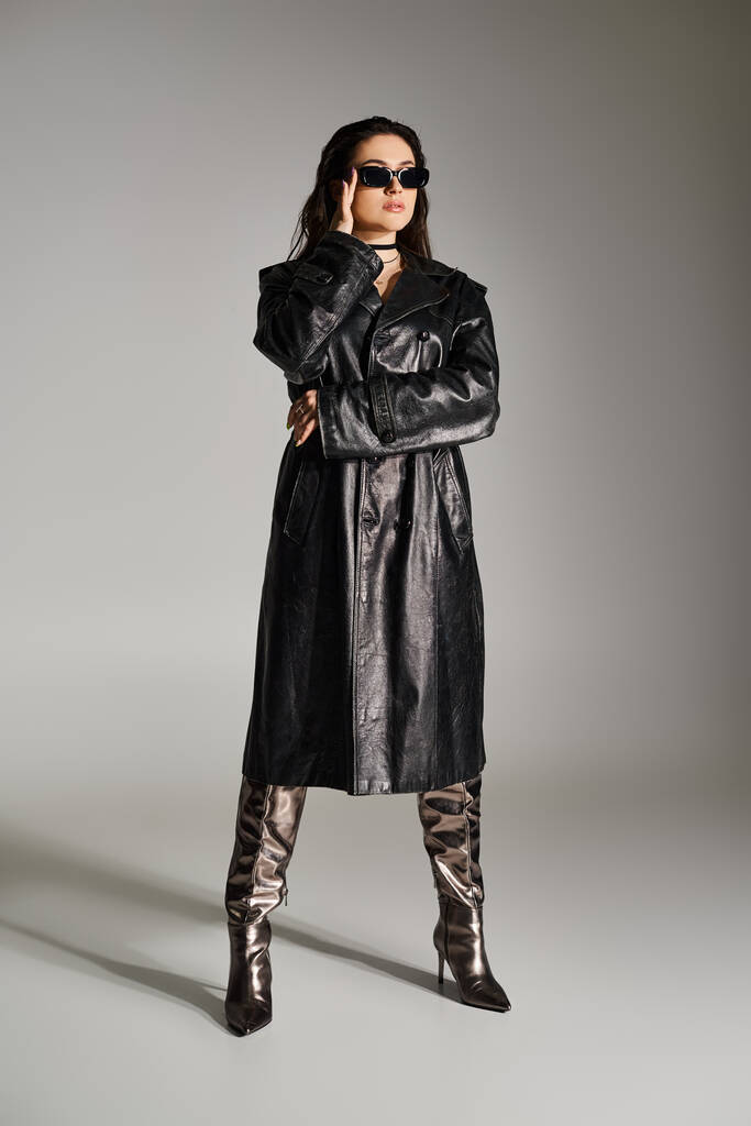 Una donna plus size trasuda fiducia in un elegante cappotto di pelle nera e stivali su uno sfondo grigio. - Foto, immagini