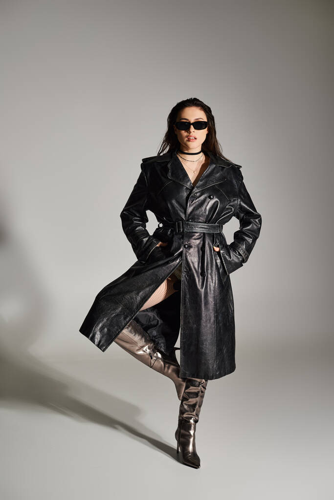 Stílusos plusz méretű nő fekete kabátban és napszemüveg pózol magabiztosan egy szürke háttér. - Fotó, kép