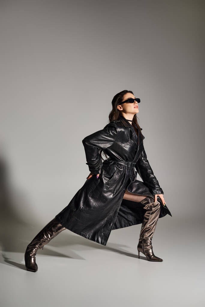 Eine schöne Plus-Size-Frau posiert selbstbewusst in schwarzem Mantel und Stiefeln vor grauem Hintergrund und strahlt Stil und Anmut aus. - Foto, Bild