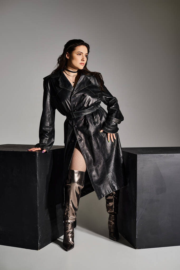 Een prachtige plus-size vrouw showcasing haar stijl in een zwarte trench jas en laarzen tegen een grijze achtergrond. - Foto, afbeelding