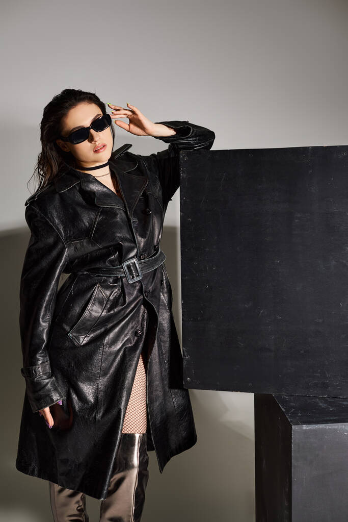 Uma mulher deslumbrante plus size faz uma pose em um casaco de couro preto e botas de coxa em um pano de fundo cinza. - Foto, Imagem