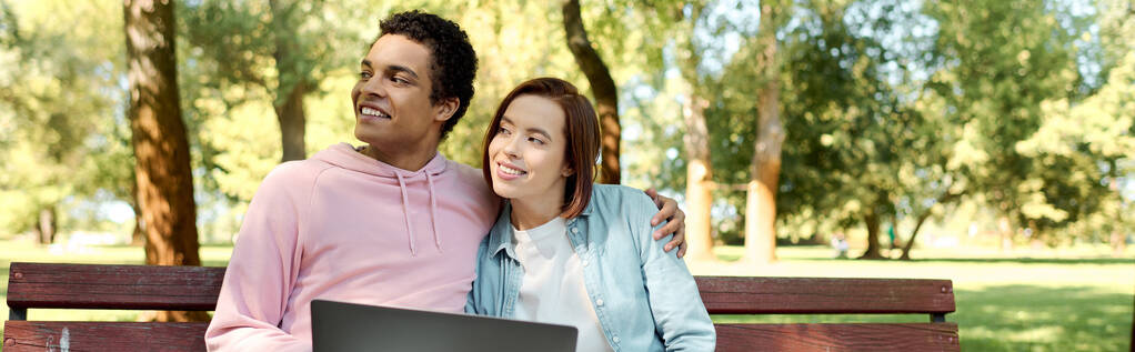 Um homem e uma mulher de traje vibrante sentam-se em um banco do parque, absorvidos em um laptop, desfrutando de uma data digital. - Foto, Imagem