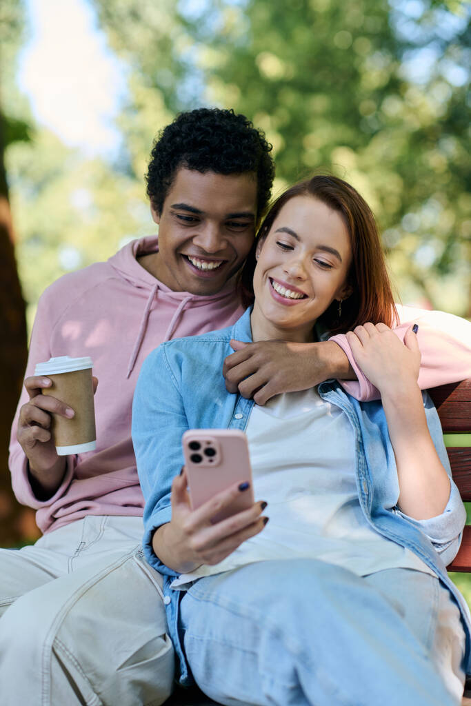 Diversa pareja en traje vibrante sentado en el banco del parque, absorto en la pantalla del teléfono celular. - Foto, imagen