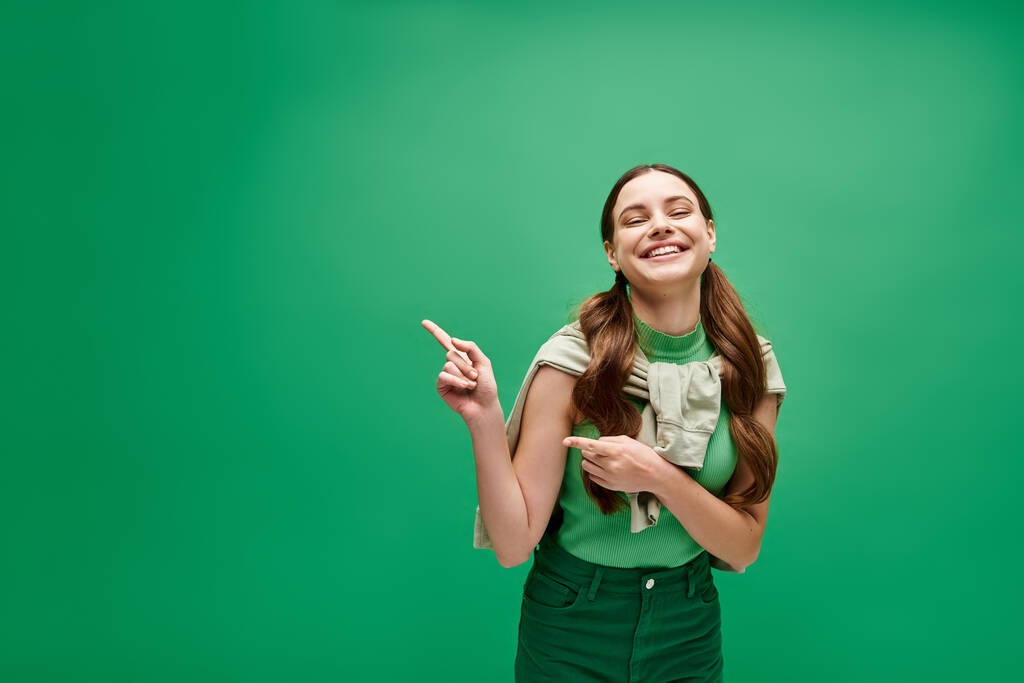 Una giovane donna sulla ventina sorride luminosa mentre indica eccitata qualcosa fuori dalla telecamera in un ambiente da studio con uno sfondo verde. - Foto, immagini