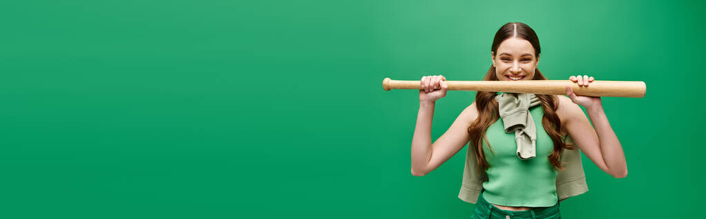 Μια νέα, όμορφη γυναίκα γύρω στα 20 κρατά ένα ρόπαλο του μπέιζμπολ μπροστά στο πρόσωπό της σε ένα στούντιο στο πράσινο.. - Φωτογραφία, εικόνα