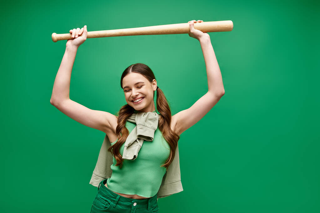 A young, beautiful woman in her 20s poses in a studio setting, defiantly holding a baseball bat over her head. - Photo, Image