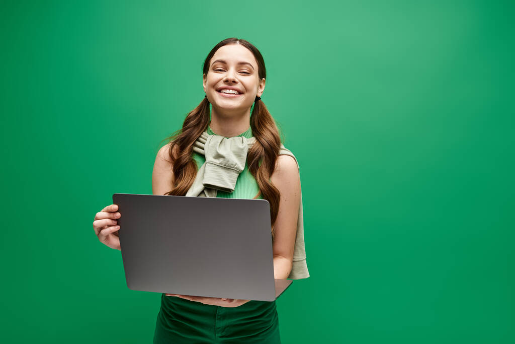 20 'li yaşlarda bir kadın canlı yeşil bir arkaplan önünde dizüstü bilgisayar tutuyor, dijital güç ve bağlantı yayıyor.. - Fotoğraf, Görsel