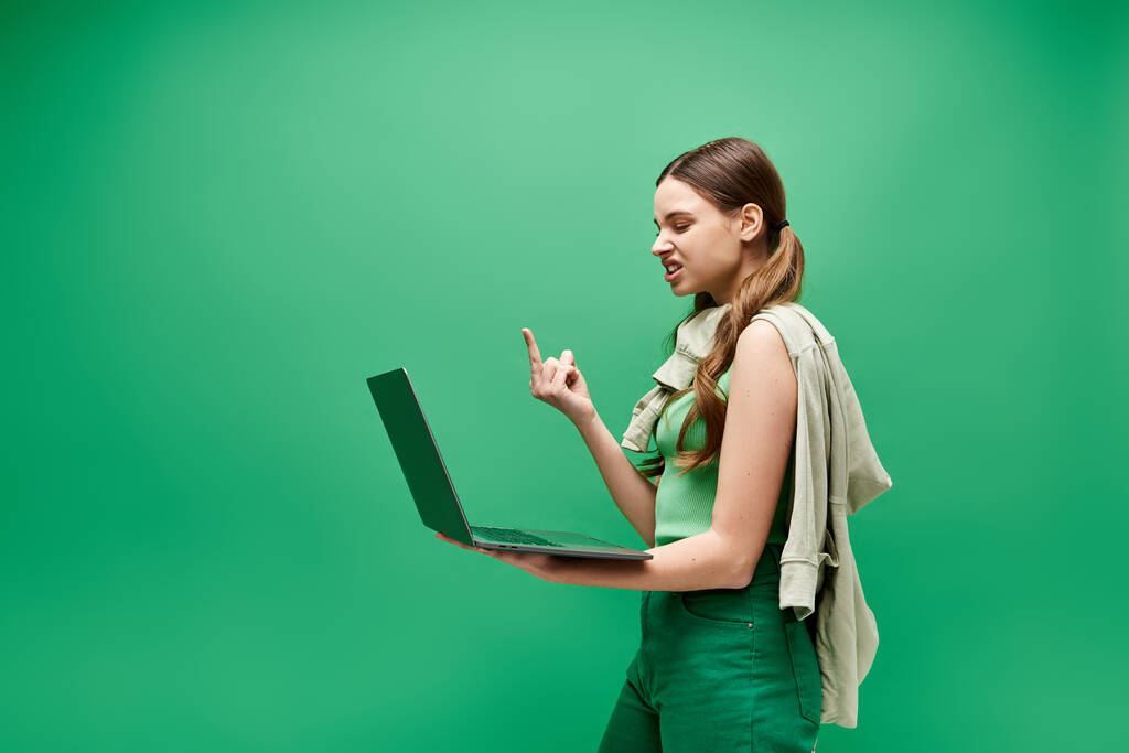 A young woman in her 20s, wearing a green attire, holds a laptop and showing middle finger in a studio setting. - Photo, Image