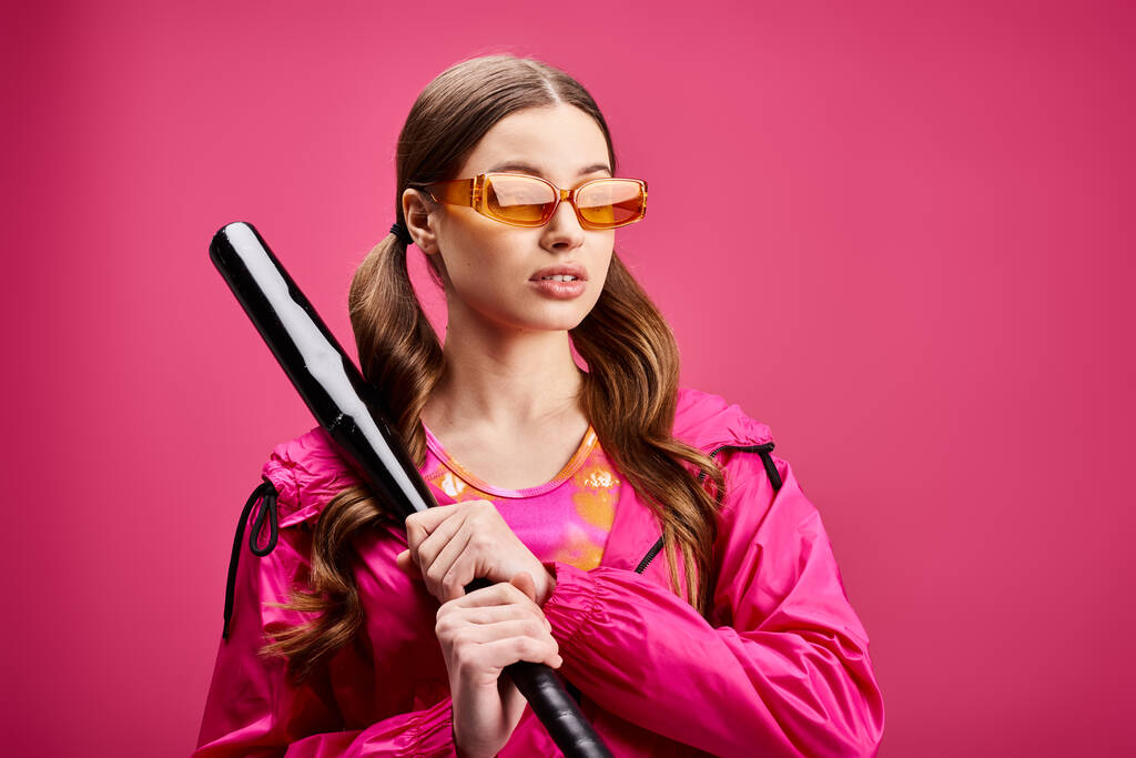 A stylish woman in her 20s, wearing a pink jacket, confidently holds a baseball bat against a vibrant pink background. - Photo, Image