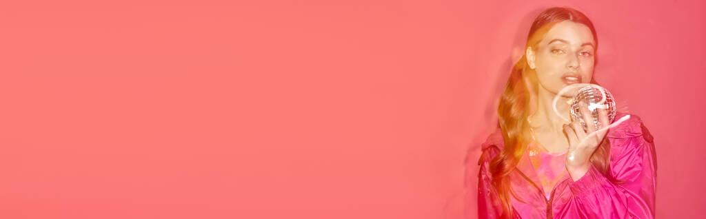 Молодая женщина в розовом платье элегантно держит диско-шар, создавая мечтательную и волшебную атмосферу в студии с розовым фоном. - Фото, изображение