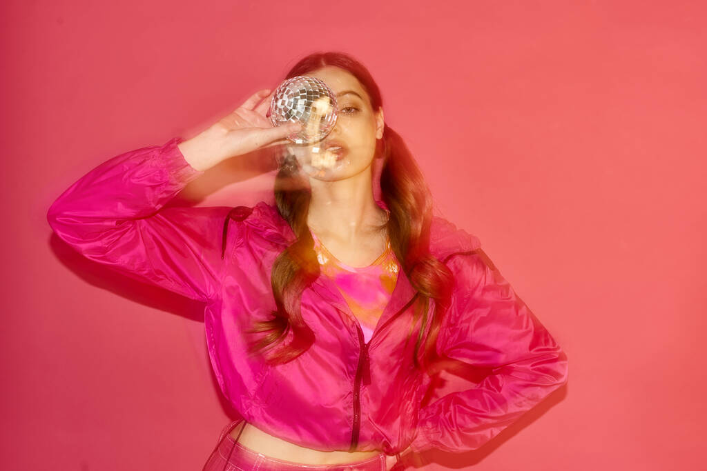 Молодая женщина 20 лет, в стильной розовой куртке, держит диско-мяч в студии. - Фото, изображение