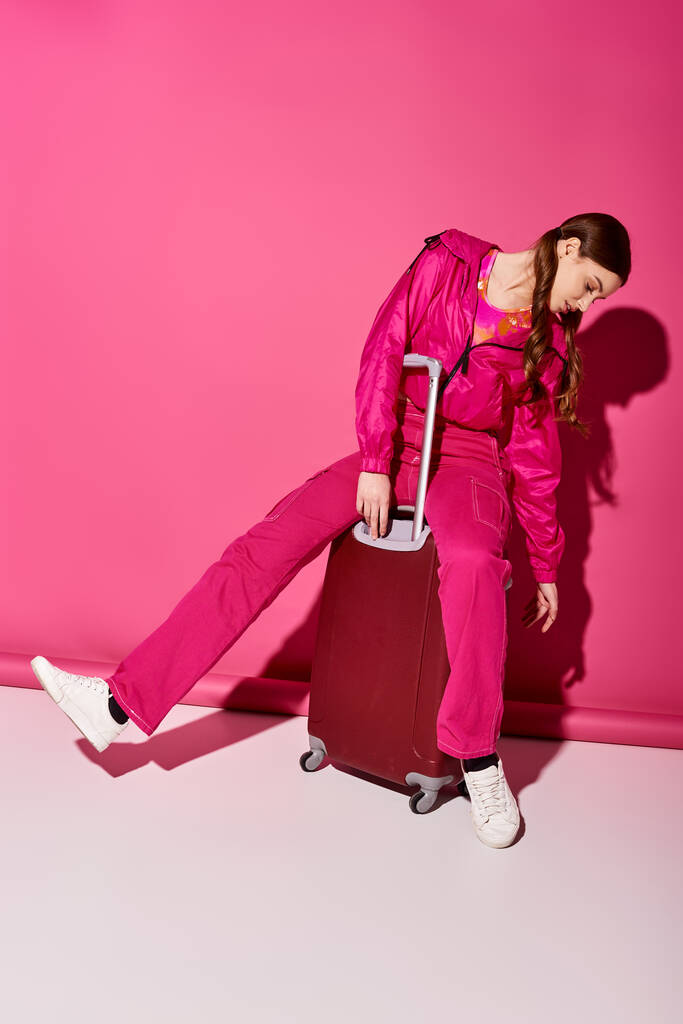 Μια κομψή νεαρή γυναίκα γύρω στα 20 κάθεται στην κορυφή μιας αποσκευής, ενσαρκώνοντας την προσδοκία για την επόμενη περιπέτεια σε ένα στούντιο με ροζ φόντο.. - Φωτογραφία, εικόνα