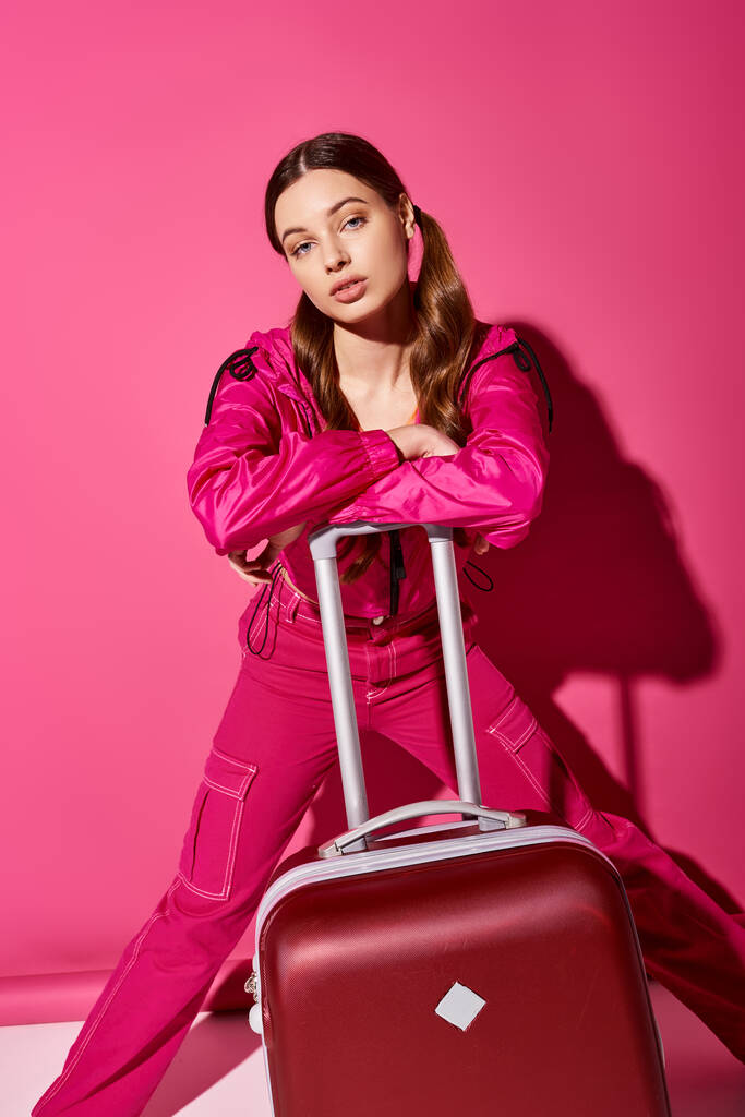 Стильная 20-летняя женщина позирует с чемоданом у яркой розовой стены, излучая элегантность и страсть к путешествиям. - Фото, изображение