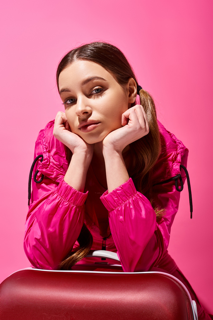 Una mujer joven y elegante de unos 20 años se sienta frente a una vibrante maleta roja contra un fondo rosa, perdido en el pensamiento. - Foto, Imagen