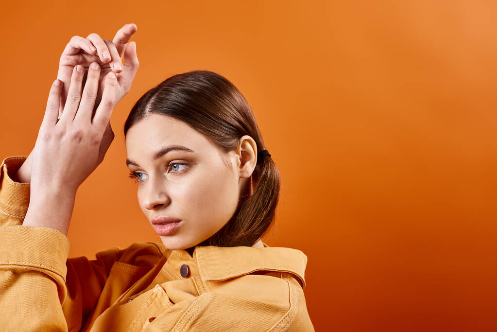 A stylish woman in her 20s raises her hands in a yellow jacket against an orange backdrop in a studio setting. - Photo, Image