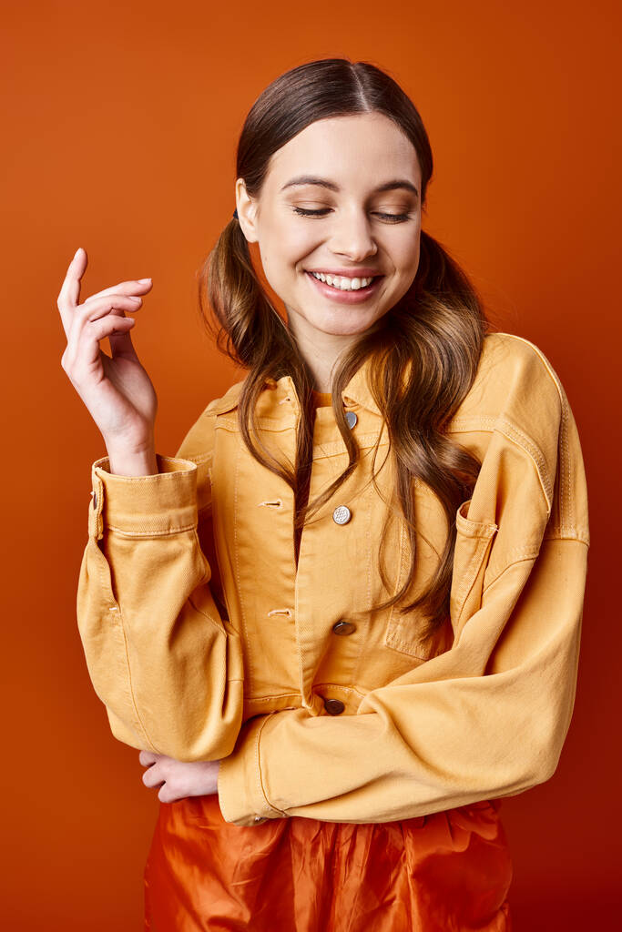 A stylish young woman in her 20s wearing a yellow shirt and pants poses in a studio with an orange background. - Photo, Image