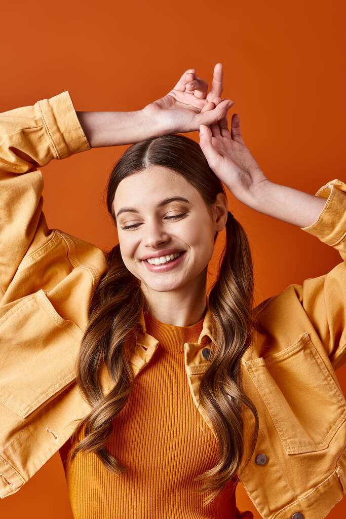 Uma jovem mulher elegante na casa dos 20 anos com uma camisa amarela, alegremente levantando as mãos acima da cabeça contra um fundo laranja. - Foto, Imagem