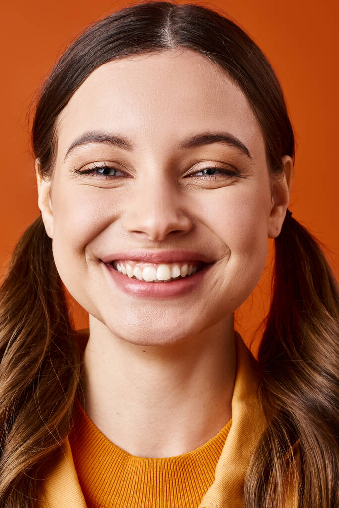 Een stijlvolle jonge vrouw van in de twintig met lang haar glimlacht fel, draagt een levendige gele trui tegen een oranje achtergrond. - Foto, afbeelding