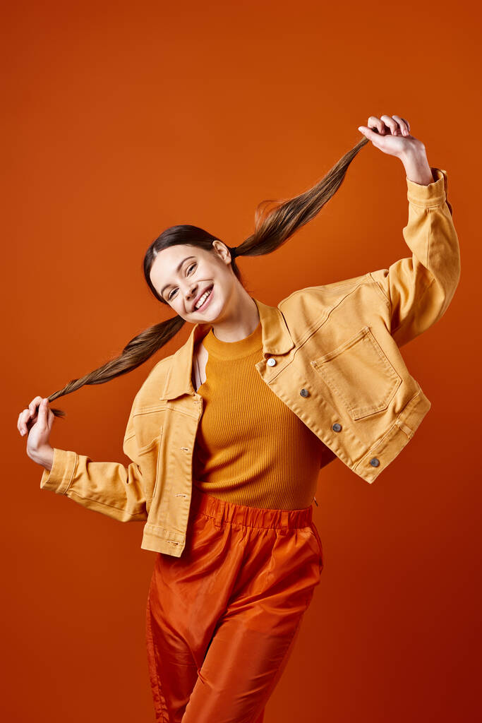Μια κομψή νεαρή γυναίκα γύρω στα 20 φοράει κίτρινο μπουφάν και παντελόνι, ποζάροντας πάνω σε πορτοκαλί φόντο σε ένα σκηνικό στούντιο.. - Φωτογραφία, εικόνα