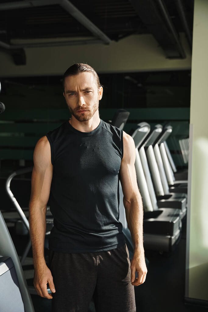 Ein athletischer Mann in aktiver Kleidung steht selbstbewusst vor einer Reihe von Laufbändern, bereit für eine herausfordernde Trainingseinheit. - Foto, Bild