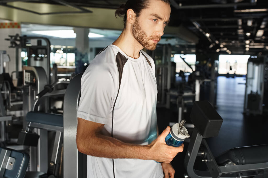Ein athletischer Mann in aktiver Kleidung macht eine Pause und hält eine Flasche Wasser in einem Fitnessstudio, umgeben von Trainingsgeräten. - Foto, Bild