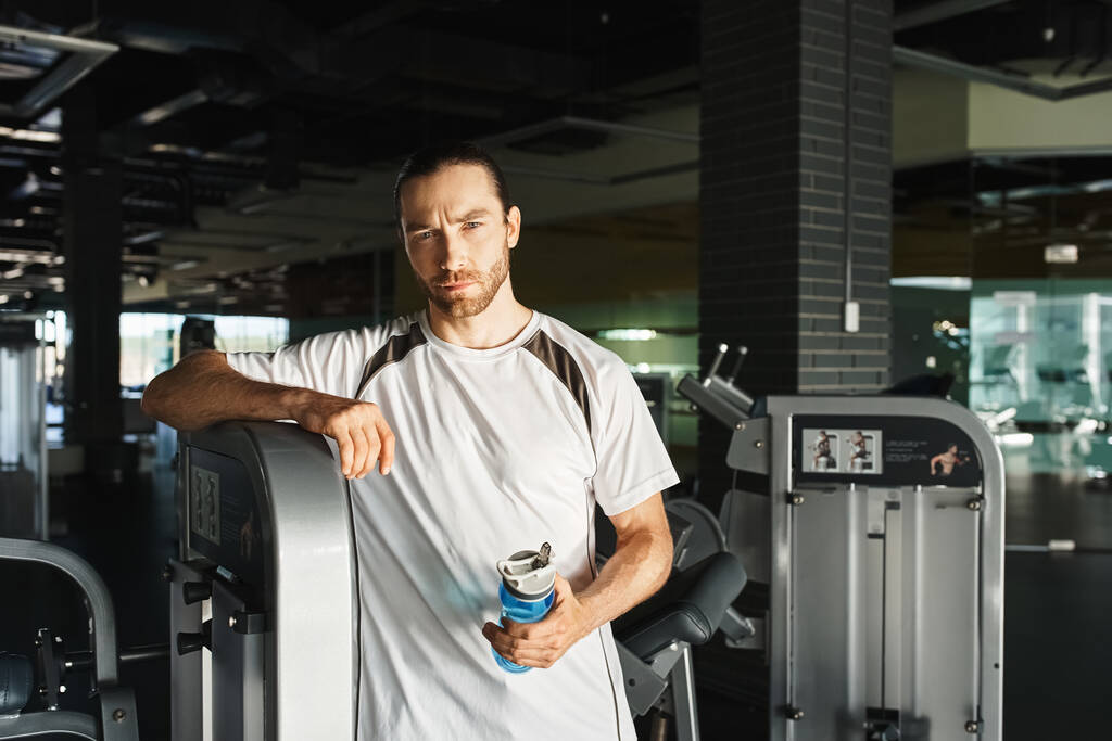Een atleet in actieve kleding staat naast een machine in een fitnessruimte en bereidt zich voor op zijn workout routine.. - Foto, afbeelding