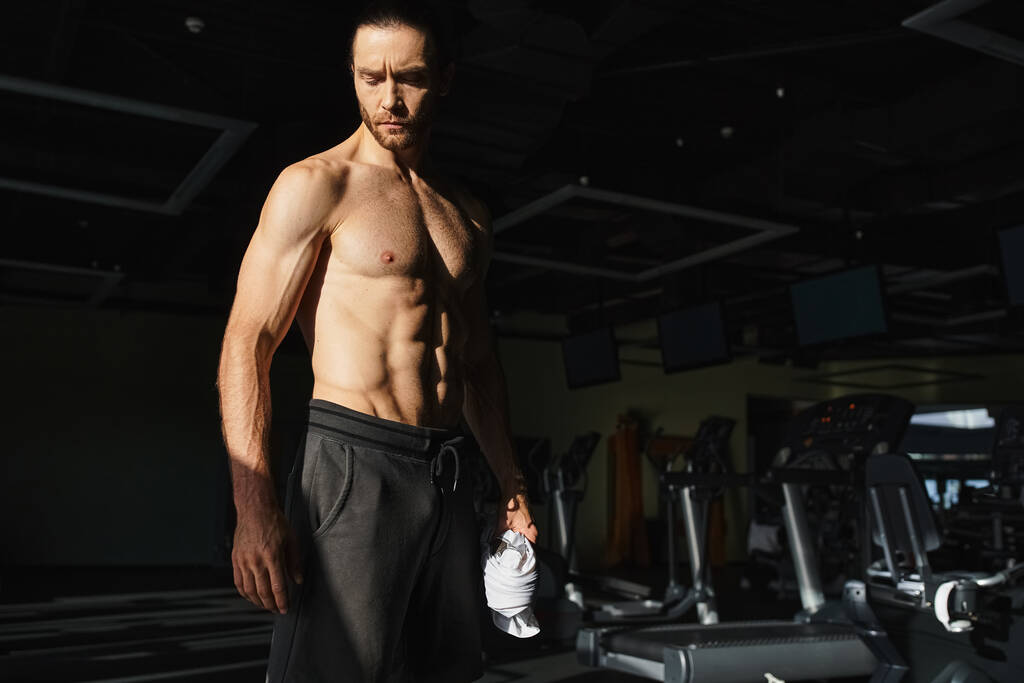 Ένας μυώδης άντρας, χωρίς μπλούζα, στέκεται σε ένα γυμναστήριο, επιδεικνύοντας τη φυσική του δύναμη και την αφοσίωσή του στη γυμναστική.. - Φωτογραφία, εικόνα