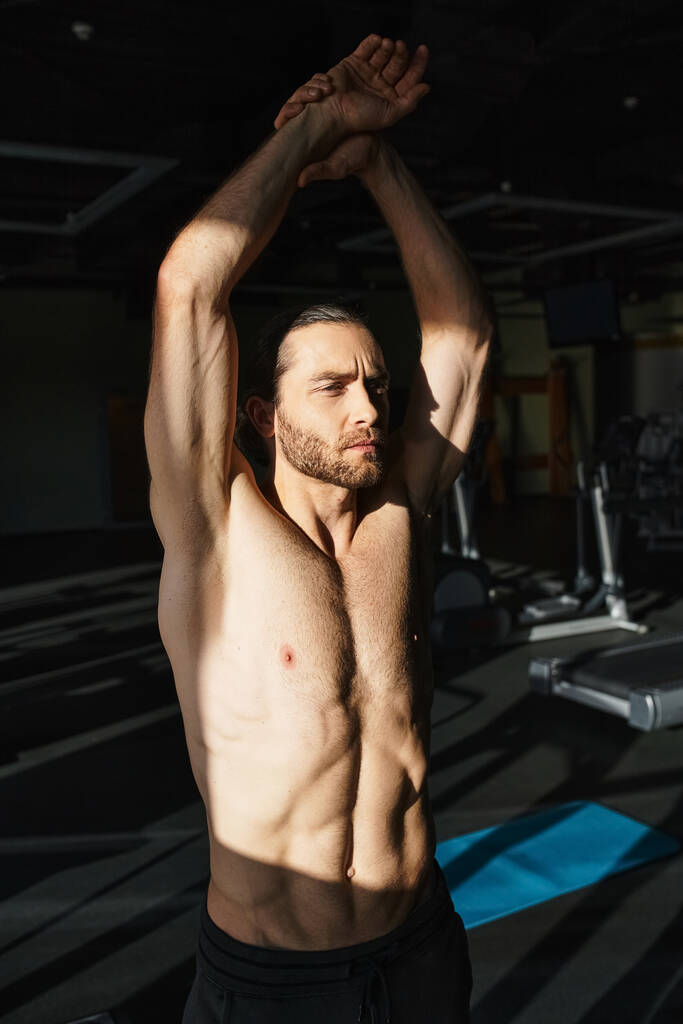 Hombre musculoso sin camisa de pie en el gimnasio con las manos arriba, mostrando fuerza y determinación en la sesión de entrenamiento. - Foto, imagen